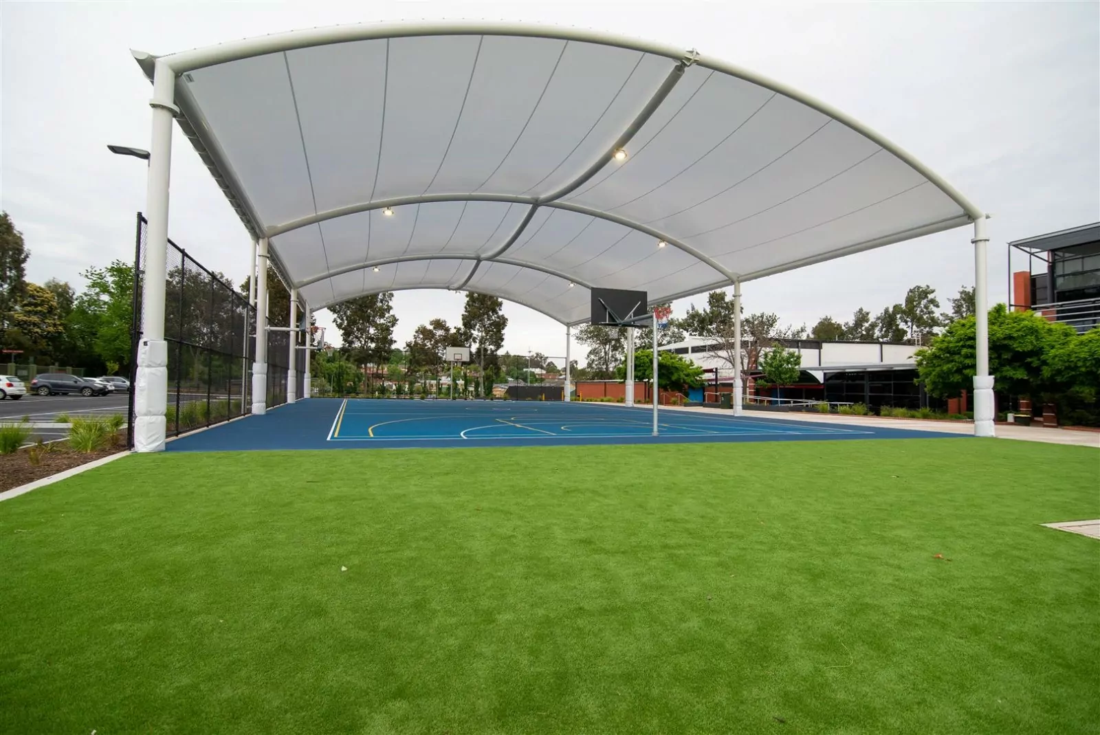 The sheltered outdoor sports court at Bendigo Senior Secondary College. The protective canopy runs the length of the blue sports court. The School's buildings stand on one side of the background and a parking lot with parked cars to the other.