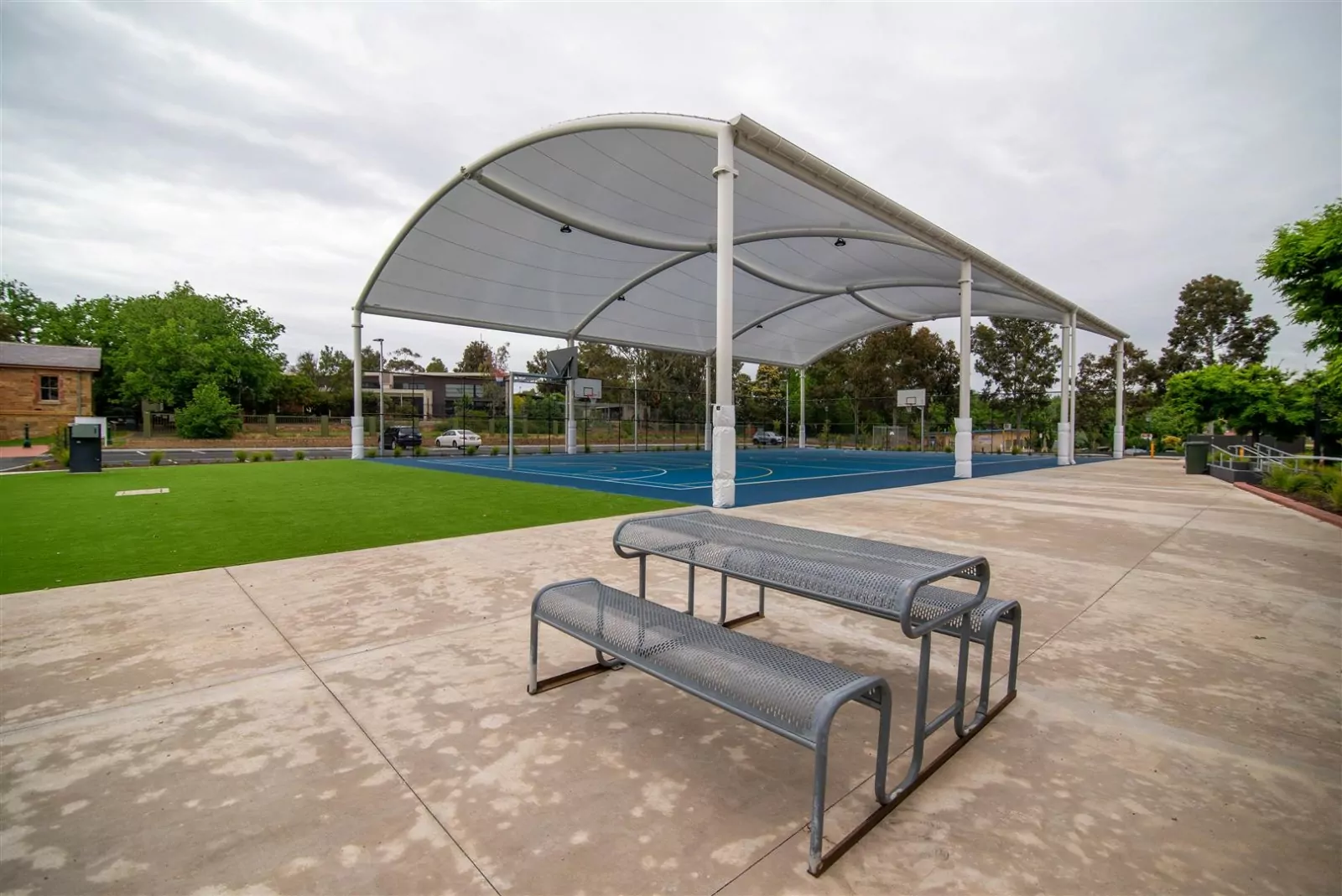 An outdoor, covered basketball court under a cloudy sky, with a metal picnic table in the foreground.