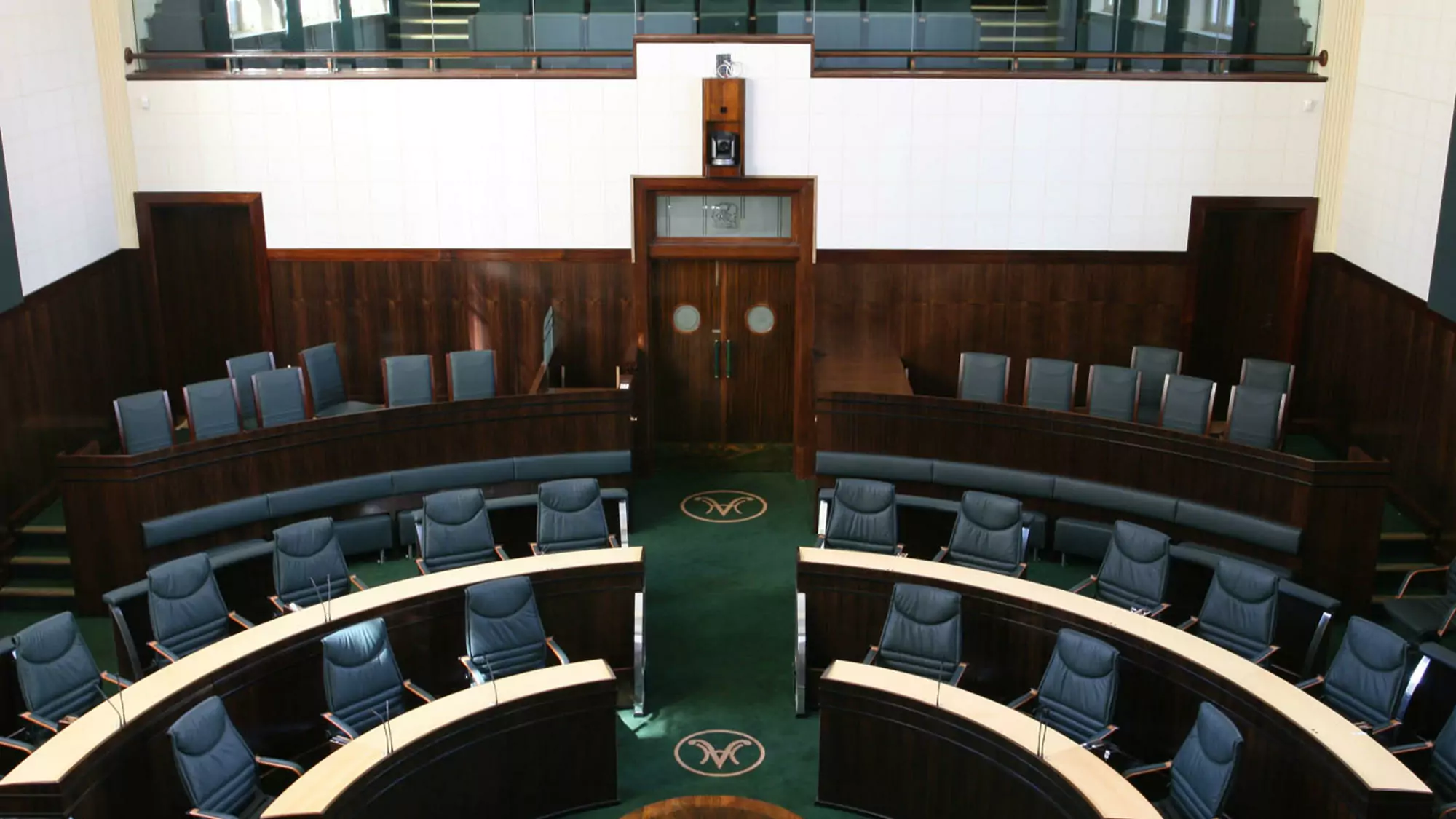 An elevated view of the House of Assembly of the Tasmanian Parliament. Curving ascending rows of green carpets, black leather seats and dark wood panels contrast the lightly painted walls.