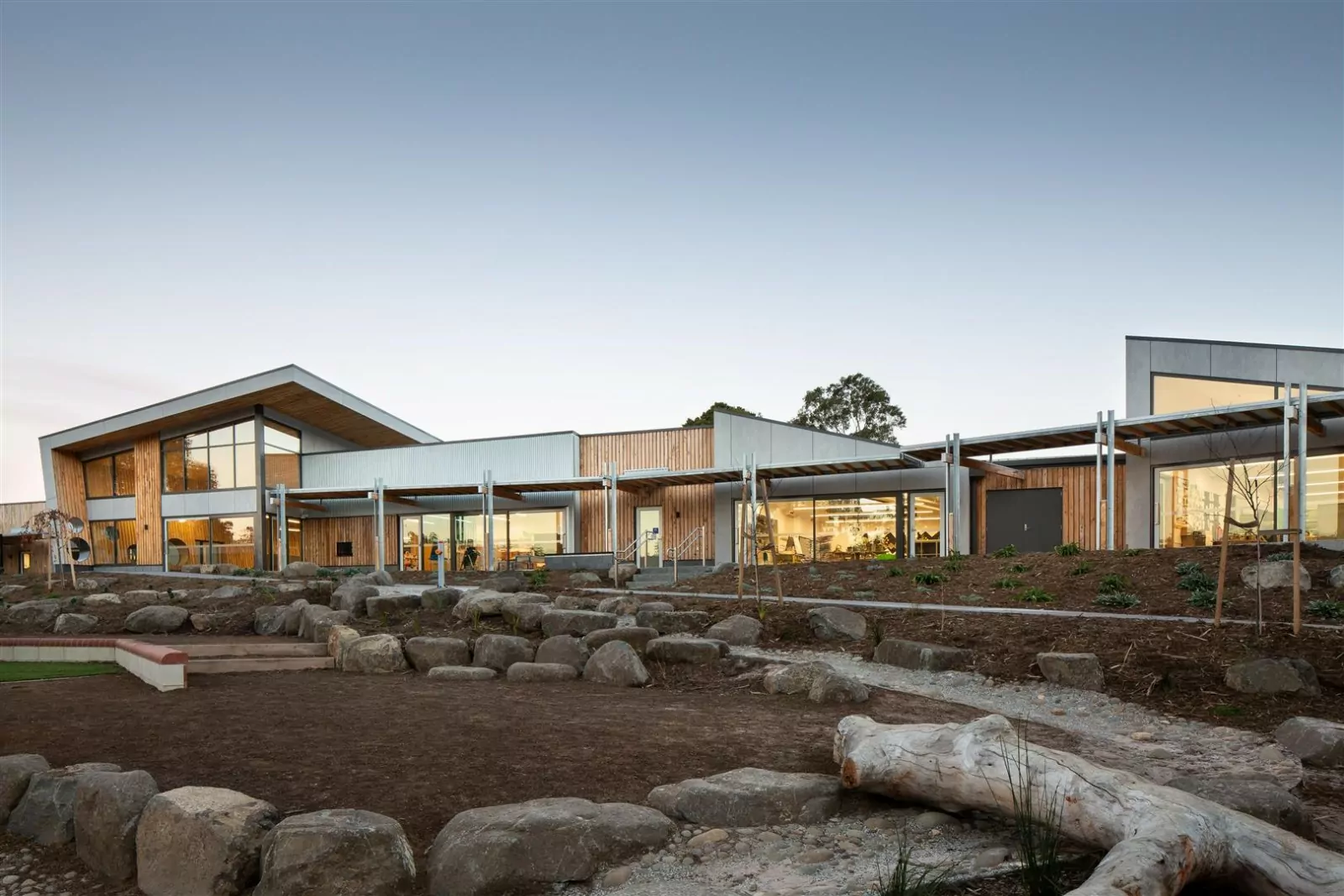 Warm sunlight reflects off the external windows of the Austin's Ferry Primary School. A walkway of small feature boulders and logs complement the wood feature walls of the building.