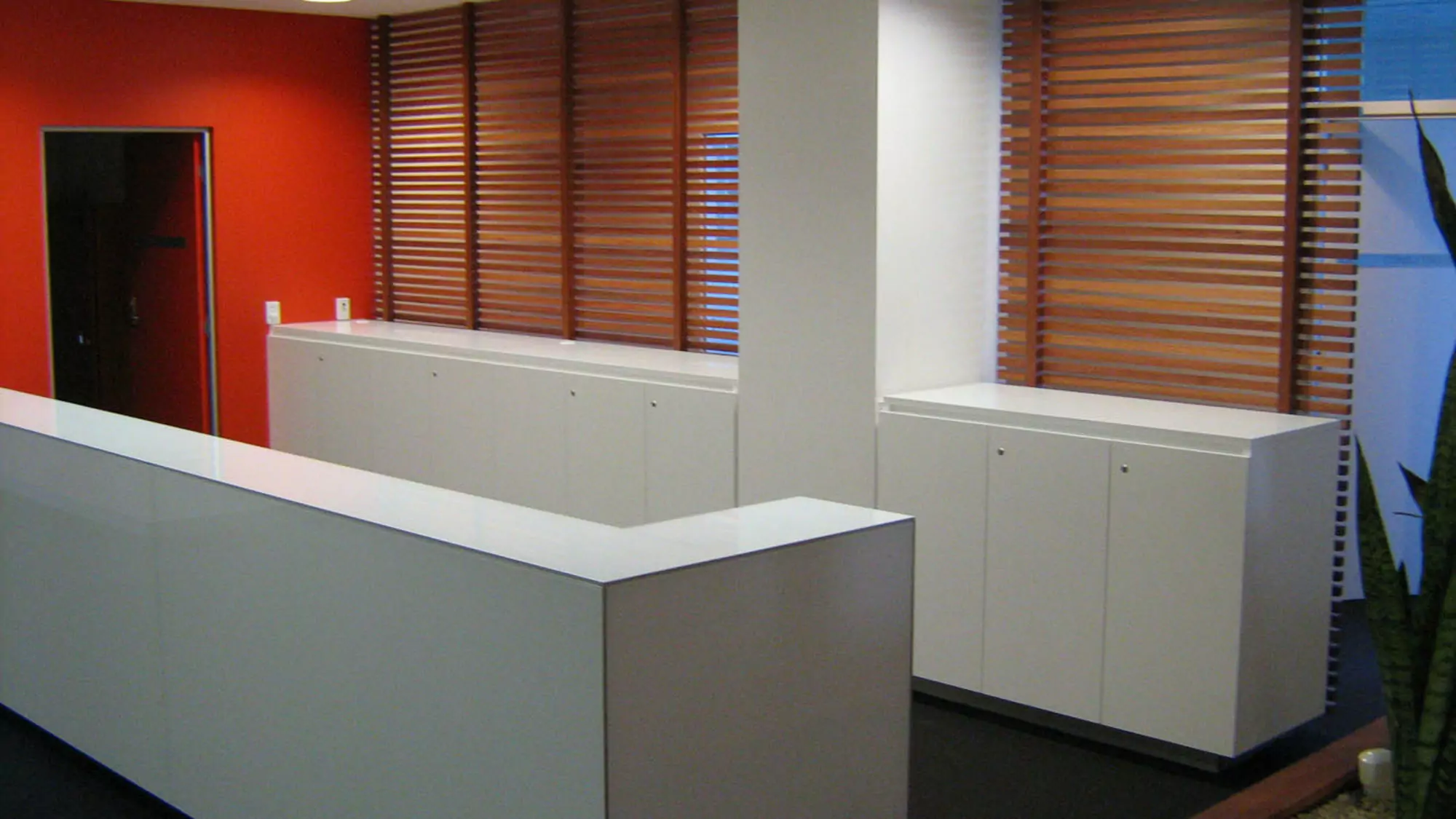 Dual rows of white cabinets lead to an open red door. Behind the further row of cabinets, thin horizontal timber beams join together to make an interior wall.