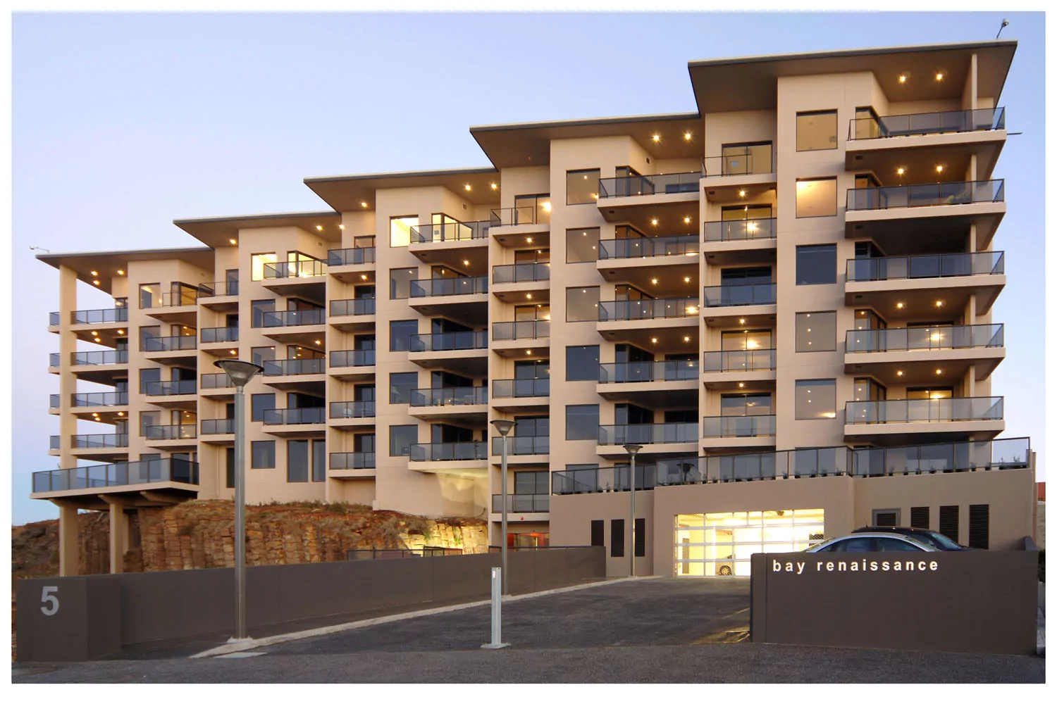 An image showing the seven-story development of 24 luxury apartments. The lights of the undercover carpark are on and there are cars parked in the outdoor carpark. The basalt rock face is visible underneath part of the building.