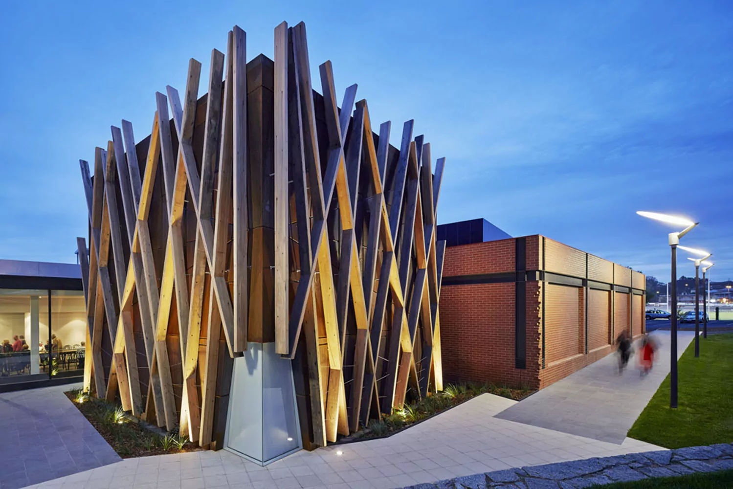 Cross, hatch-patterned timber planks run the length of a building's exterior corner. On one side of this building, two blurred out pedestrians walk past a redbrick building. To the left, a gathering of people are visible through the window.
