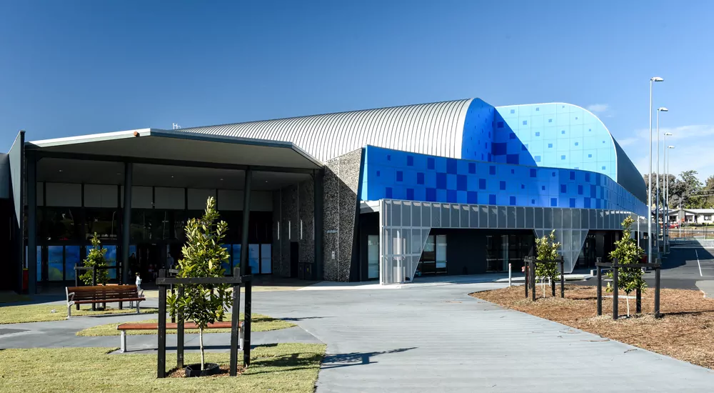 A tall, prominent structure of the Bendigo Stadium occupies much of the photo. The blue patterns on its roof match sky above. The foreground is a concrete walkway lined with small trees in the lead up to tall glass entrance windows and doors.