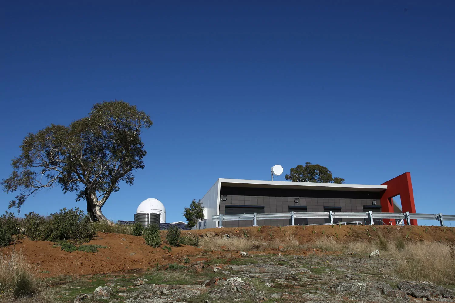 Atop a rocky hill, behind a metal road guardrail, the Observatory Dome of the Bisdee Tier Observatory stands out against the clear blue skies. The adjacent administration building has white Kingspan sandwich panel cladding and roofing with a bright red feature wall.
