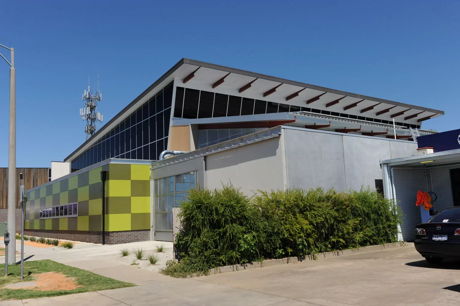The near side of a large building has two tiered walls. One with a mix of light and dark green checkered big squares. The one above is entirely covered with large solar panels. Beneath this building there is a narrow row of neatly-cut plants and high shrubbery bush fence.