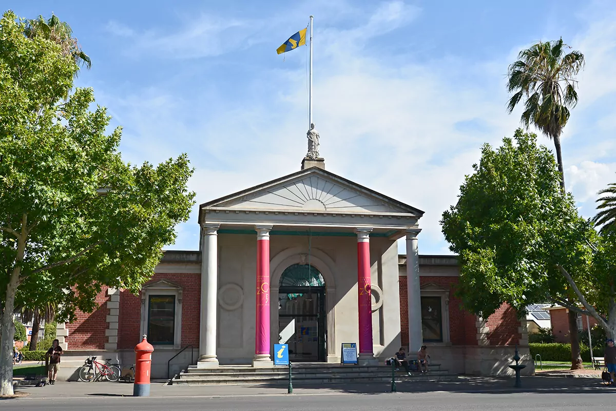 A street-side view of the neo-classically designed Castlemaine Market Building. The steps leading up to the building are flanked by large green trees and four very tall Greek-stylised columns to the Castlemaine Visitor Information Centre. The statute and flag peak the photo. The building features a mixture of white concrete and red brick walls.