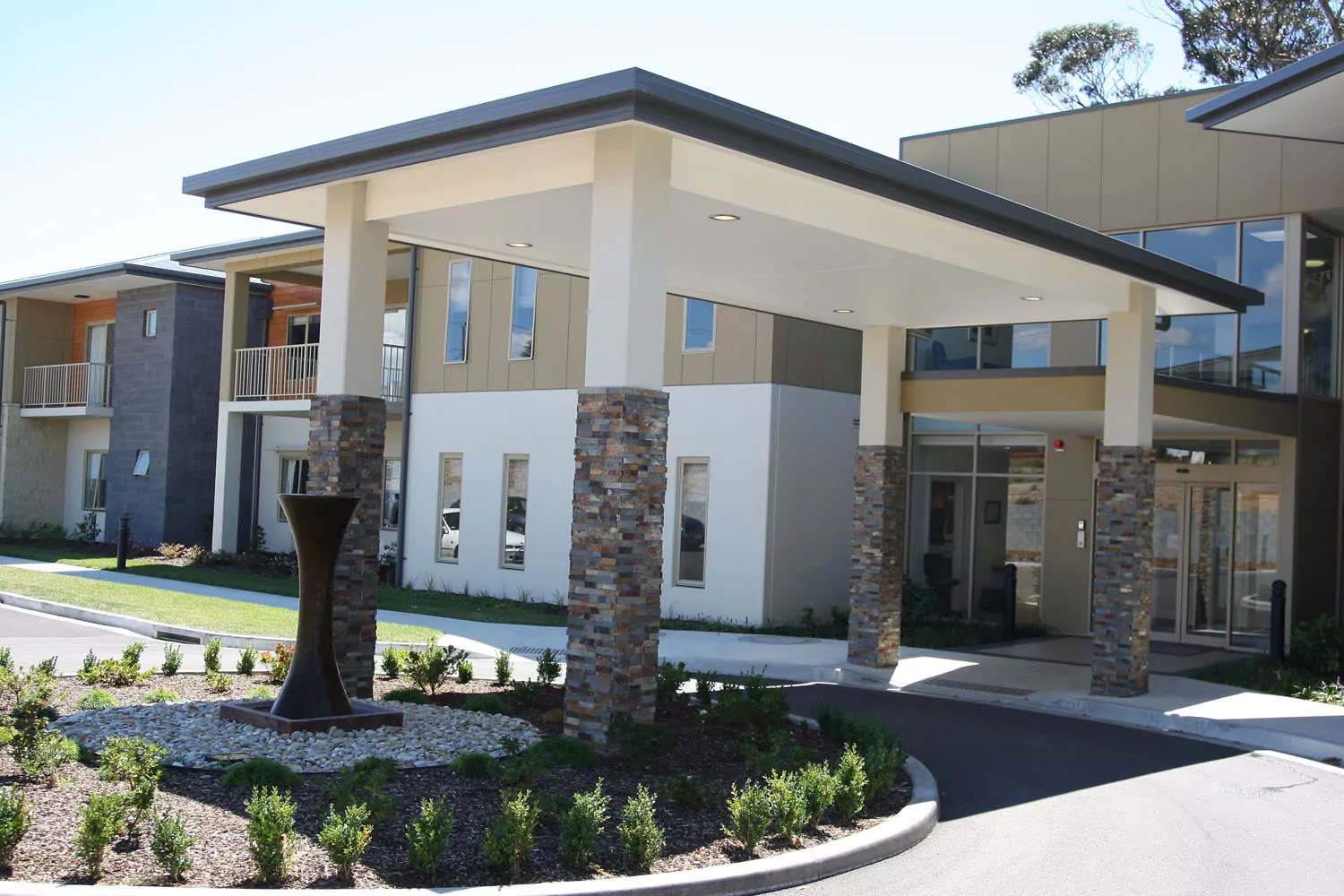 An artfully decorated roundabout connects to the entrance of a large aged care facility. Above the roundabout four pillars with stone features support a canopy.