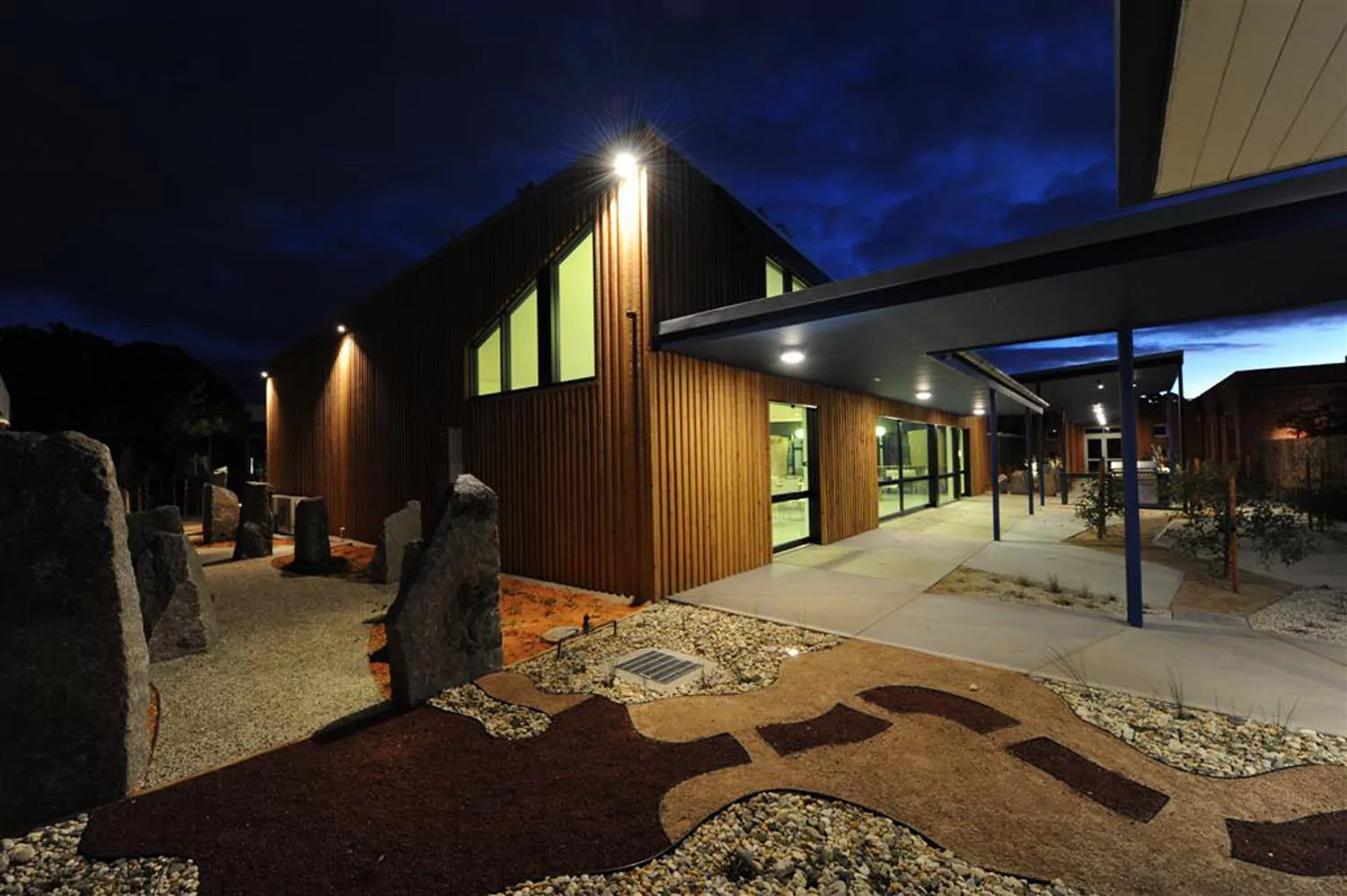 A nighttime photo of the illuminated breakout employee space for the Department of Sustainability and Environment in Epsom. The building has a timber façade. In the background there is a large open-plan modern kitchenette. The main feature of the foreground are the large granite pillar structures, colourful stones and concrete paved area in the shape of an Aboriginal possum pelt