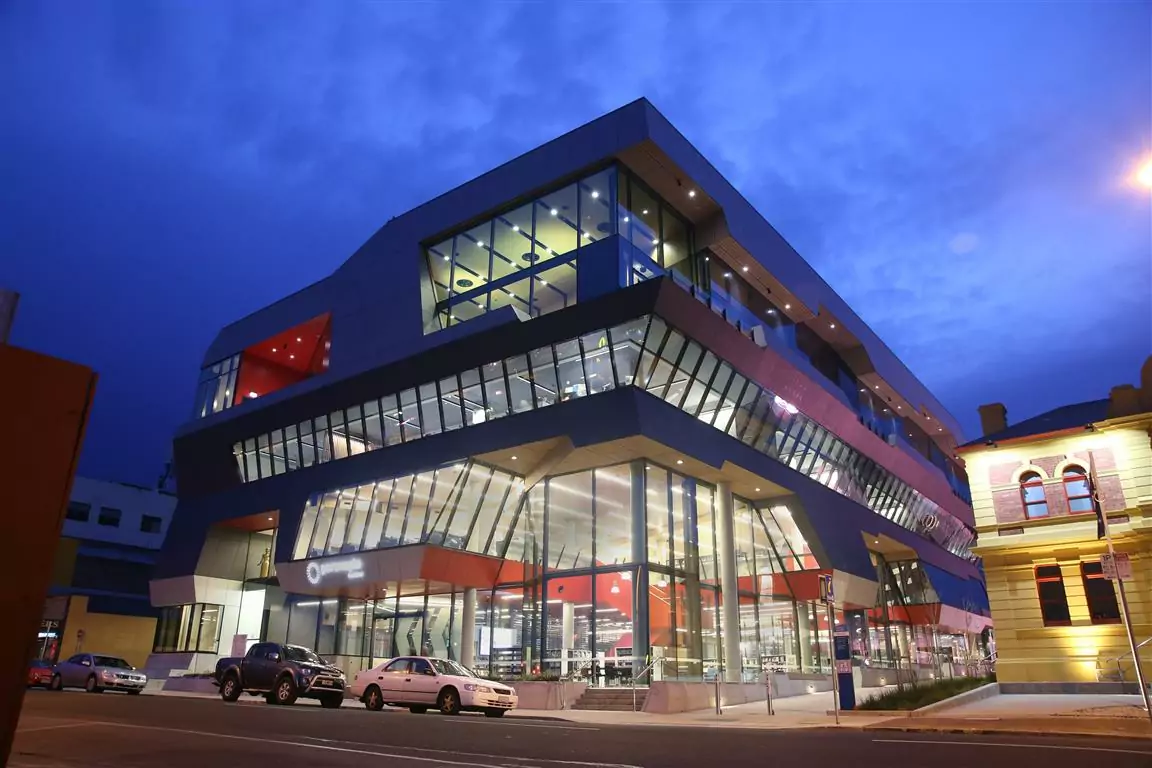 A nighttime street view of the illuminated the Paranaple Convention and Arts Centres of the Devonport Living City project with parked cars on the street curb.