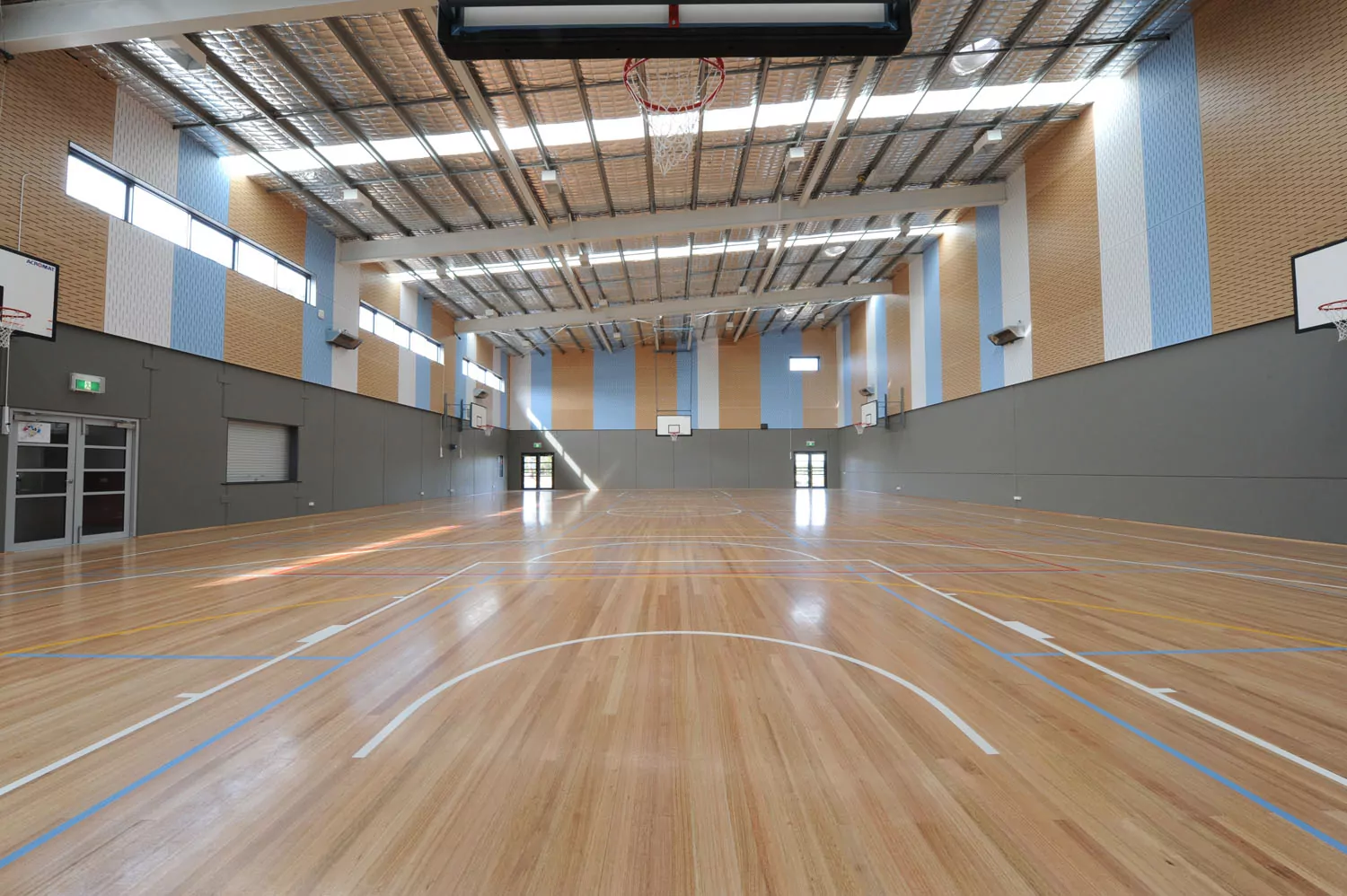 The Eaglehawk Secondary College gymnasium featuring a full-size indoor basketball court and a stage. The line markings of the court match the blue and white of the school colours, matched in bright colour streaks through the exposed precast panels and MDF lining.