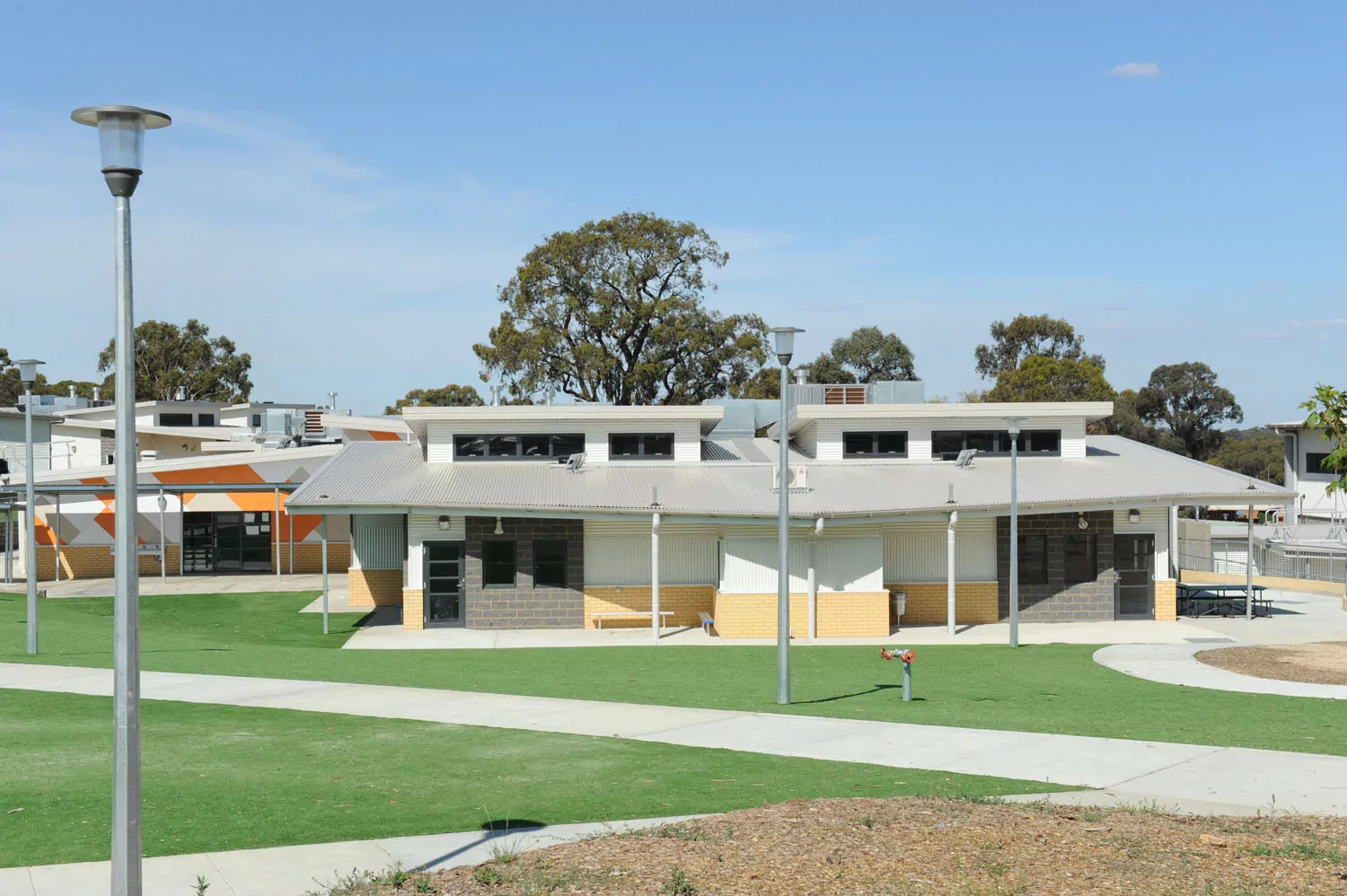 Tall lamppost fixtures stand out along the concrete path intersecting landscaped grass areas of the Eaglehawk Secondary College. A sleak profile, angular building with cream bricks and patterned walls are the central focus of the photo.