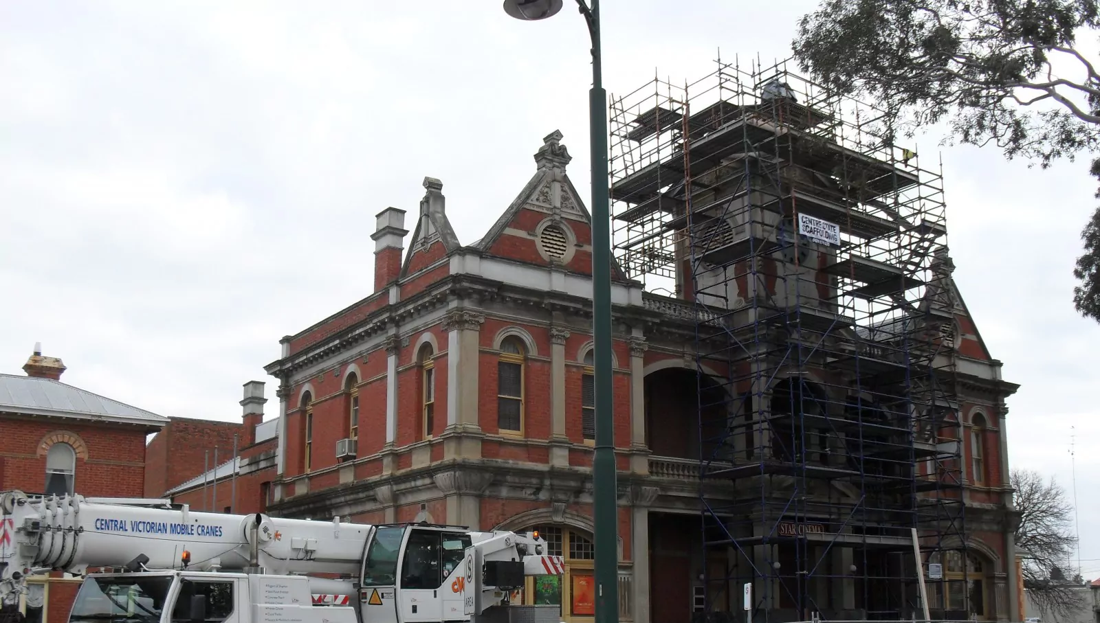 Scaffolding works to replace and repair of a section of roof and reinstatement of the flagpole on the Eaglehawk Town Hall. The community heritage building features a red brick and concrete façade. A white truck with a crane is parked on the street in front of the building.