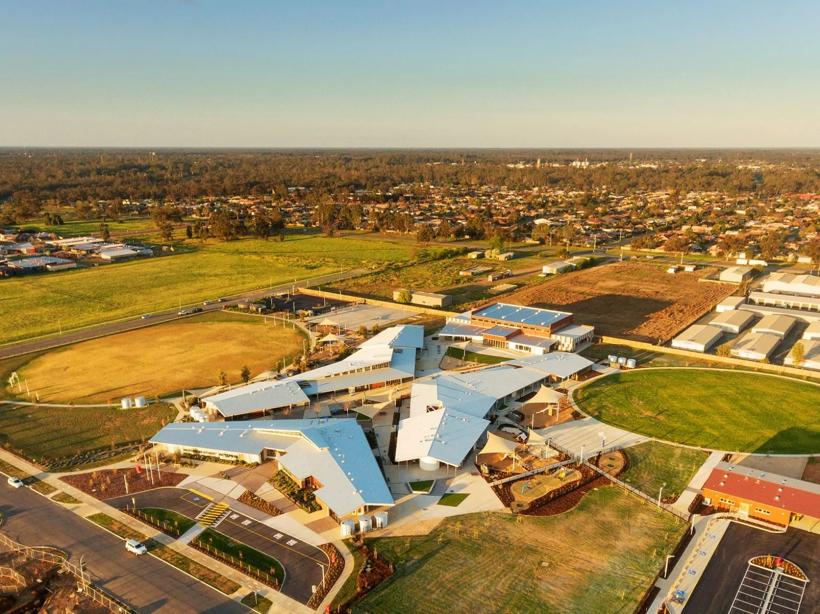 An aerial photo of the Echuca Twin Rivers Primary School. From above, there are large interestingly designed buildings that converge. Around these, there are a workshop with bus parking, gym with full-sized basketball court, kitchen stage and classrooms. Additionally, other standout features are the irrigated sports oval, two basketball courts, play equipment, lawn and garden areas, sandpits, and a paved courtyard area. A carpark for staff off-street parking.