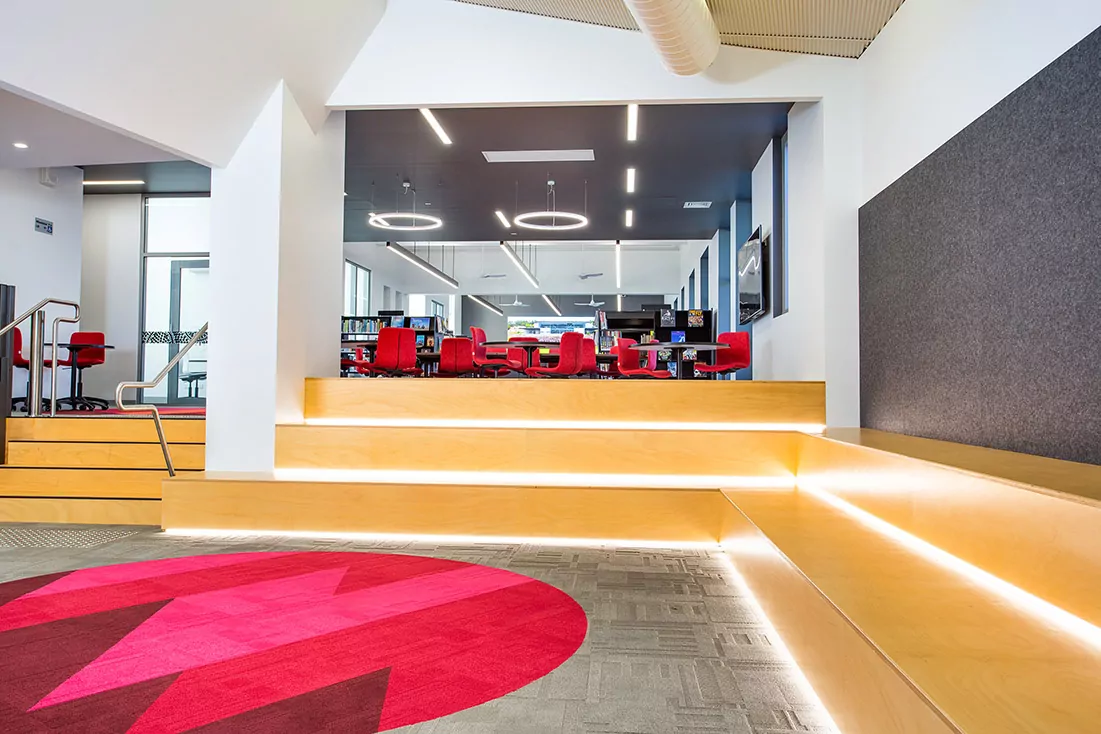 The freshly renewed, refurbished Emmanuel College Library. The grey timber floor features a red circle with internal zig-zag patterns. Behind this, a mixture of functional wooden steps and seating leads to a student area with red chairs. In the distant background, there are filled bookshelves.