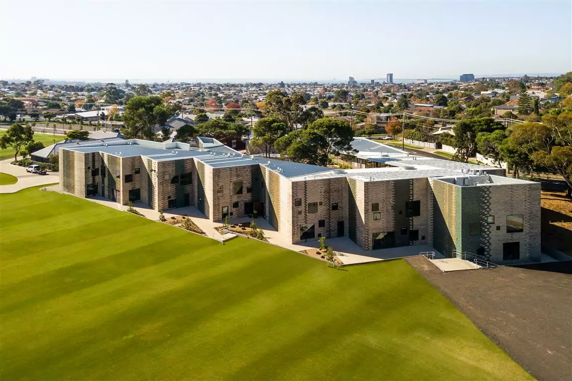 An aerial drone photo of the external facade of the Geelong College Junior School. In the foreground there are well-manicured lawns that lead to tall patterned buildings. The external areas feature a discovery garden with water pumps, creek beds and adventure trails, designed to spark interest and creative exploration with the environment.