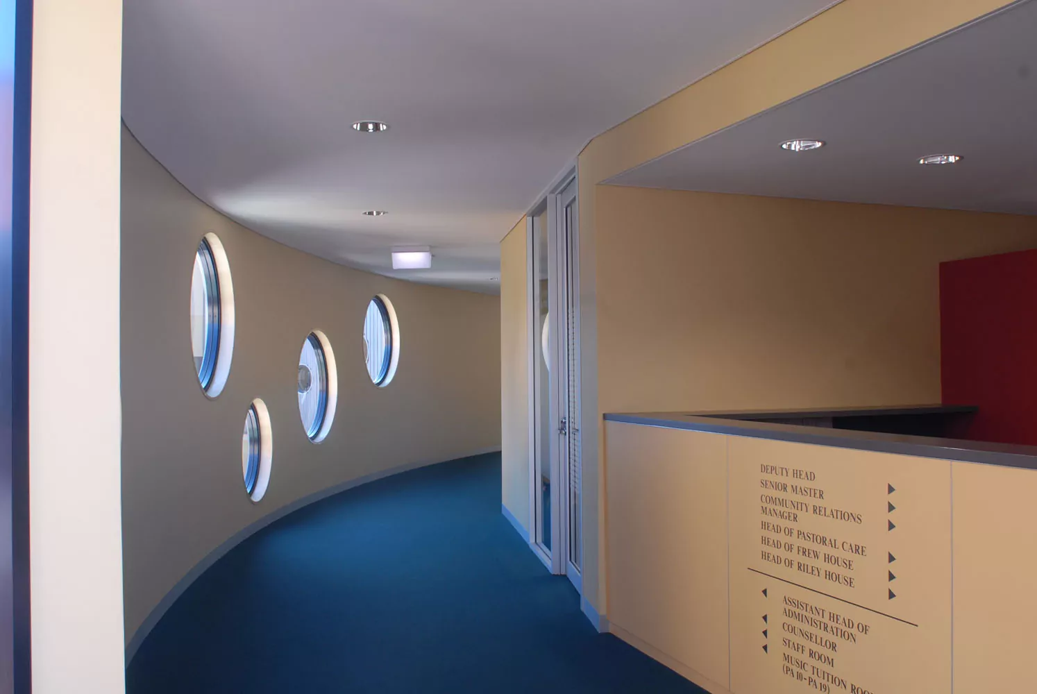 A corridor of the Girton Grammar Performing Arts building. There are blue carpeting, four circular windows and is a wall sign providing directions to various parts of the building.