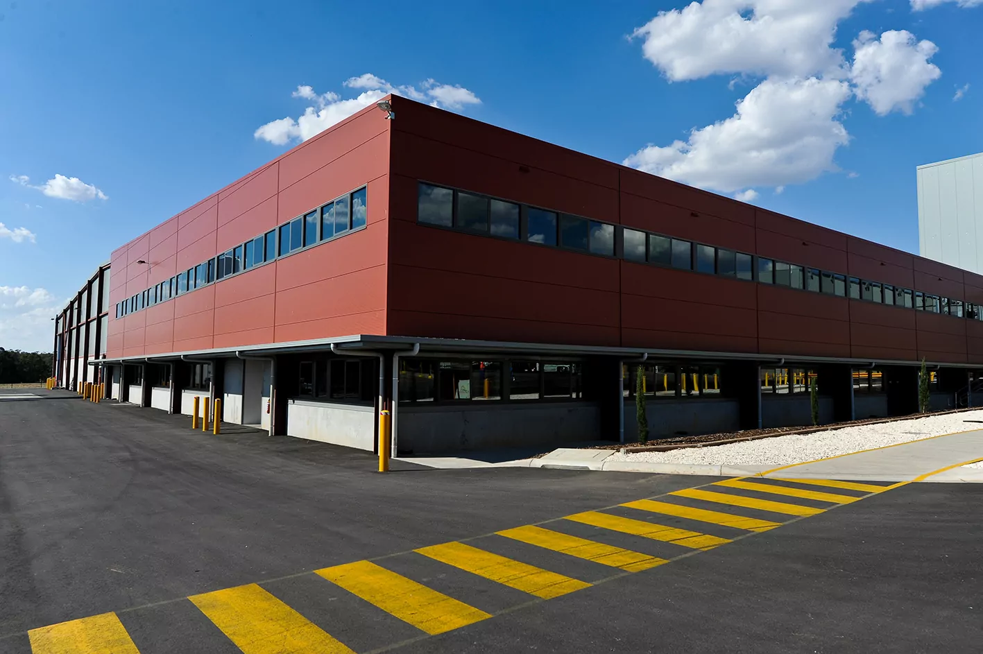 The state-of-the-art poultry processing facility for Hazeldene's Chicken Farms. A marked yellow zebra-crossing runs across a wide service road used for the delivery of poultry for processing. The large building has red Kingspan thermal cladding with windows running its length.