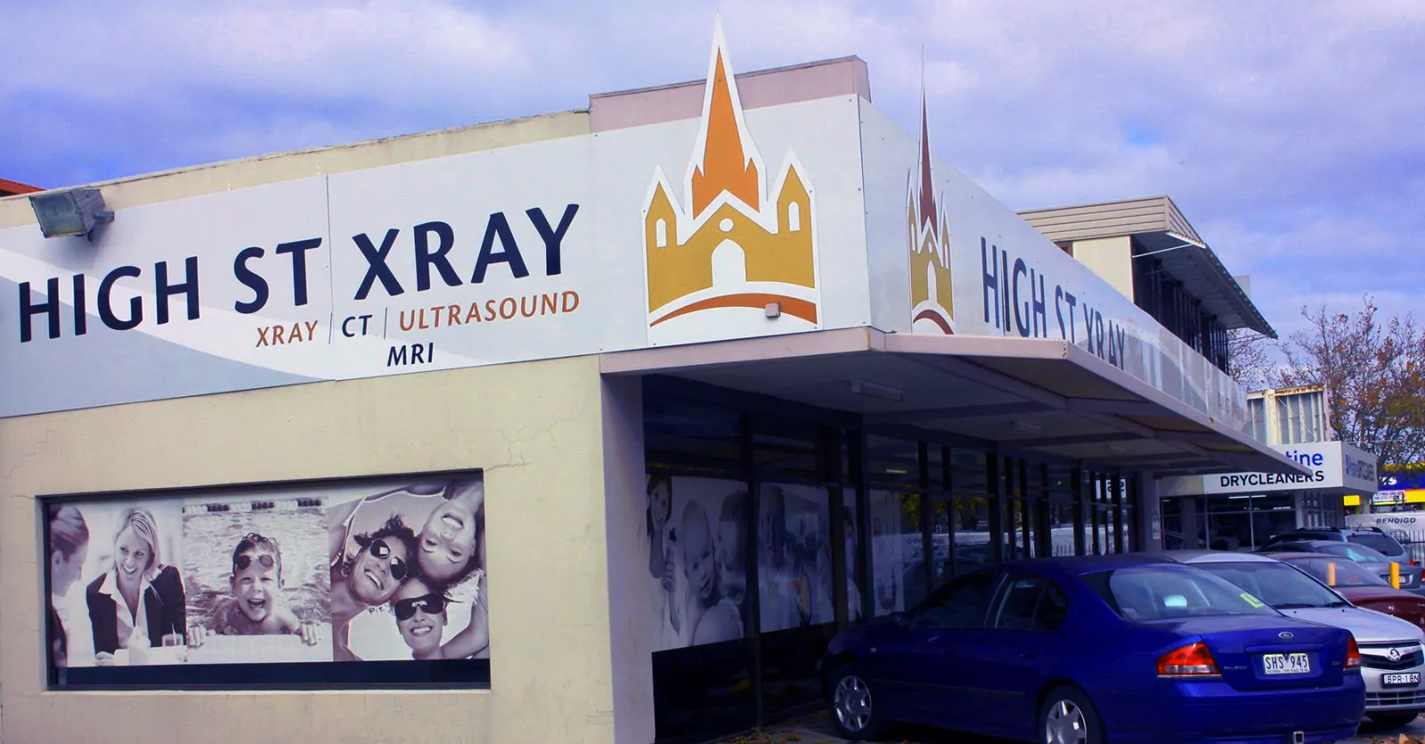 A streetview of the shopfront for High Street X-Ray. The building signage features their logo and brand name. The windows are frosted with stock photos of smiling people. Cars are parked outside the building.