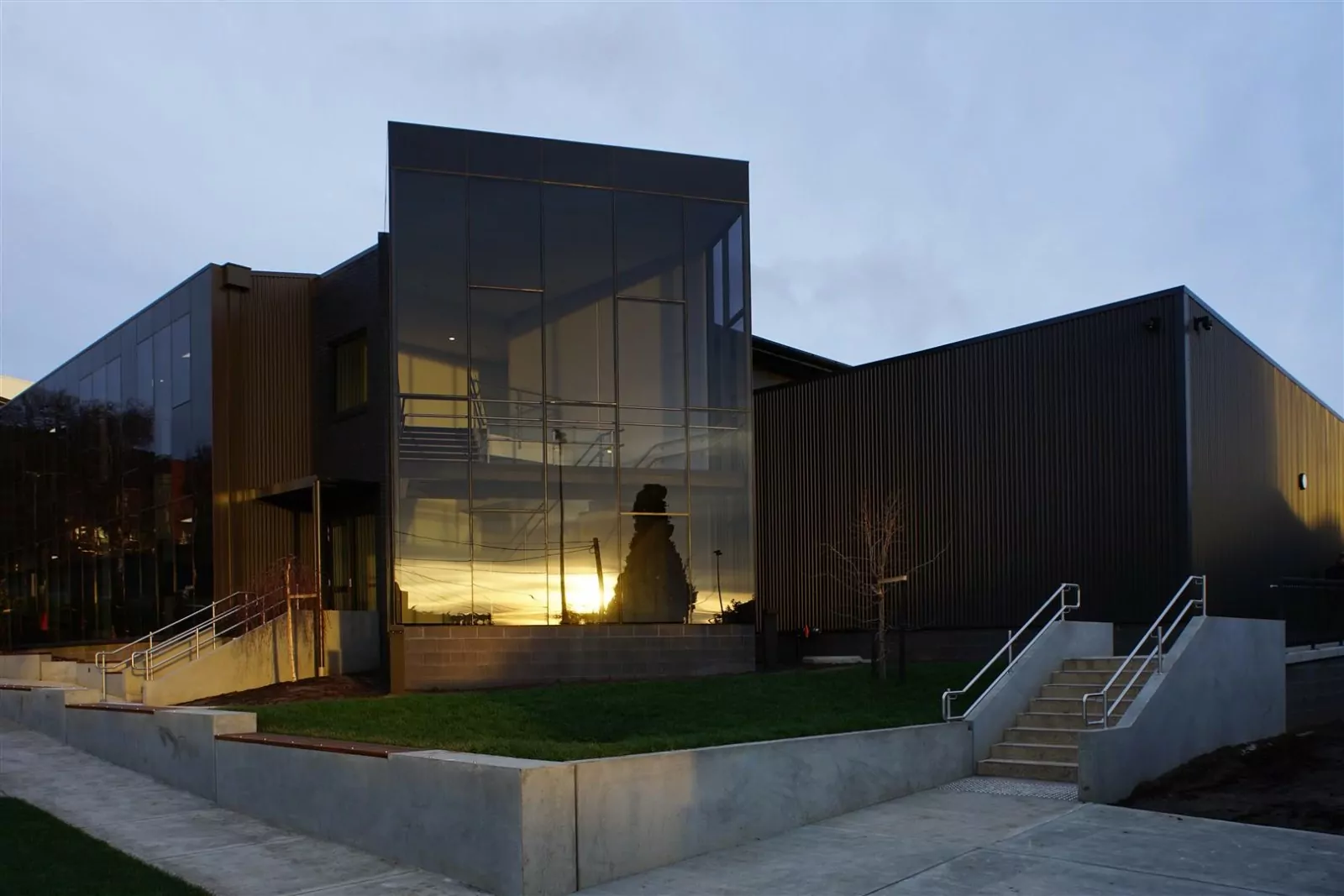 The new Chris Rae Building at the Hutchins Middle School. A sunset is reflected in the glass facade. A concrete pavement runs the perimeter of the building, intersecting a small grass section and set of stairs and