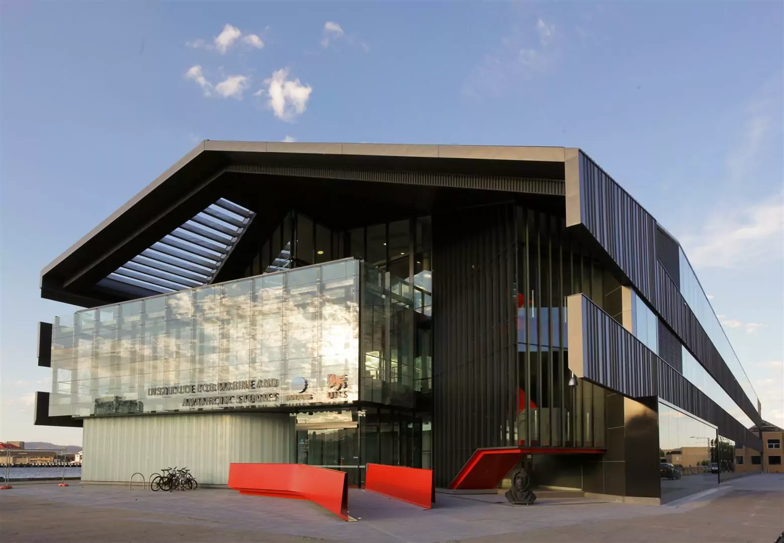 The "shed" profile of the Institute for Marine and Antarctic Studies (IMAS) on Hobart's Sullivan Cove. Sunlight and a lightly clouded sky are reflected in the building's glass. The red external features catch the viewers eye.