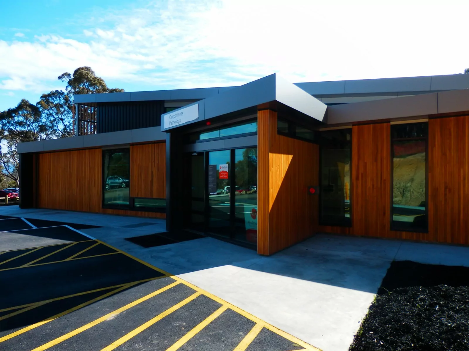 The building profile of the Kilmore and Districts Hospital casts a shadow on the surrounding parking and pedestrian walkway. Nearby parked cars and buildings are reflected in tall windows cut into wood panel walls.