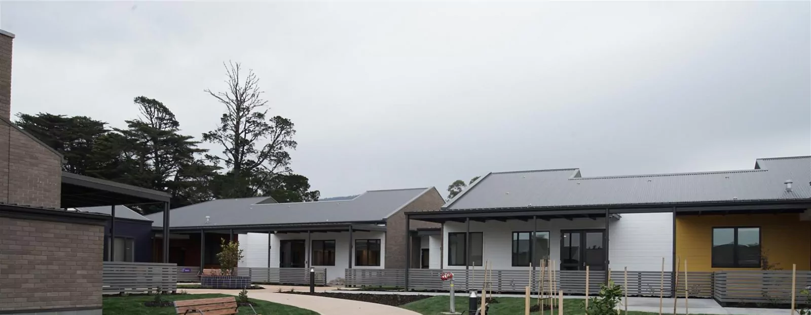 The world-class purpose built Korongee Dementia Village. An overcast sky sits above buildings designed around model small houses in the enclosed community village. The photo is taken from a pedestrian walkway with bench seating, grass areas and grey fencing around the houses.