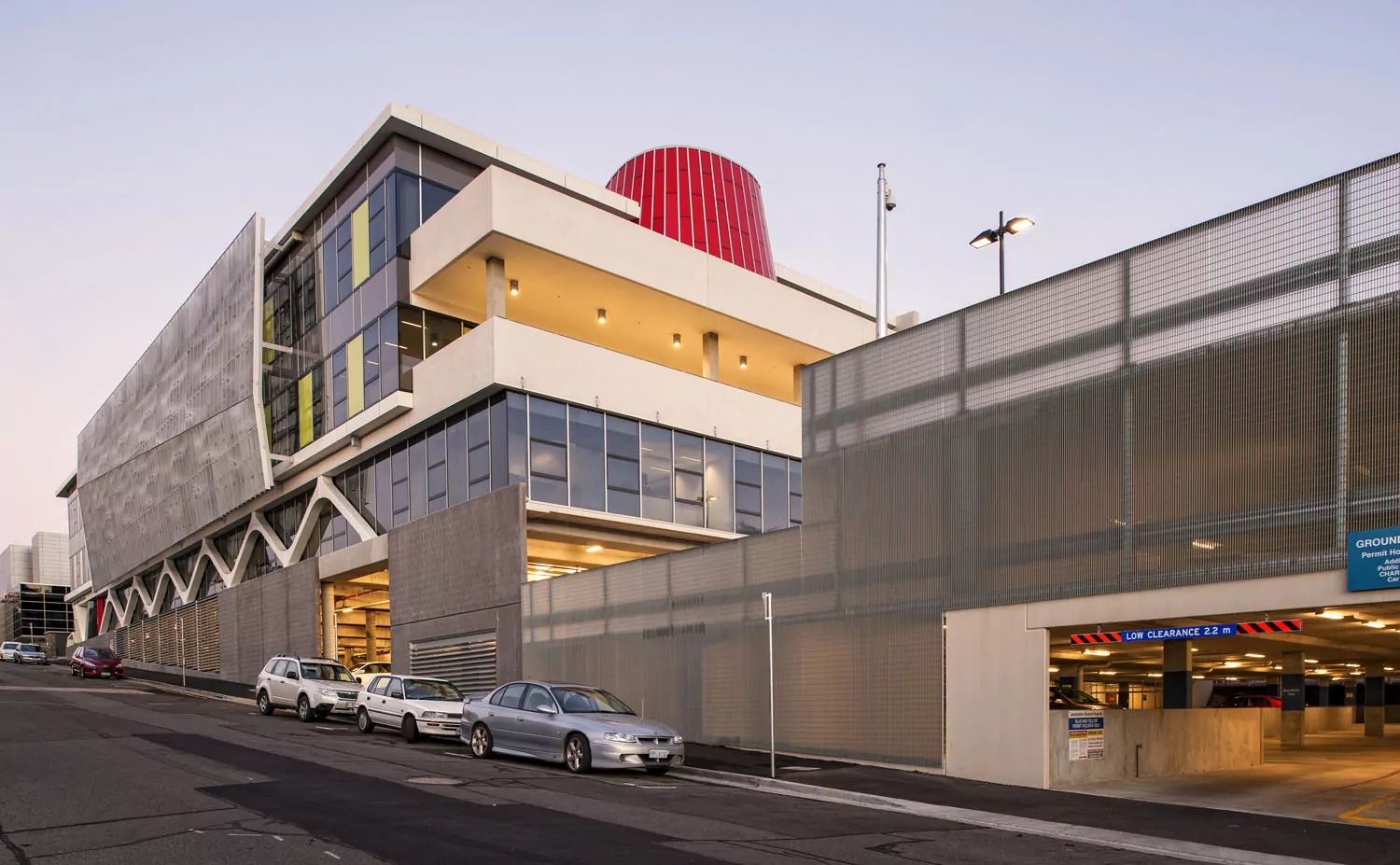 A streetview of the iconic Launceston General Hospital. A road with parked cars leads to the undercover car park There are exterior finishes of patterned pre-cast panels, pre-cast curve struts, curtain glazing and coloured glass segments. The feature red glass panels boldly constrast against the clear sky.