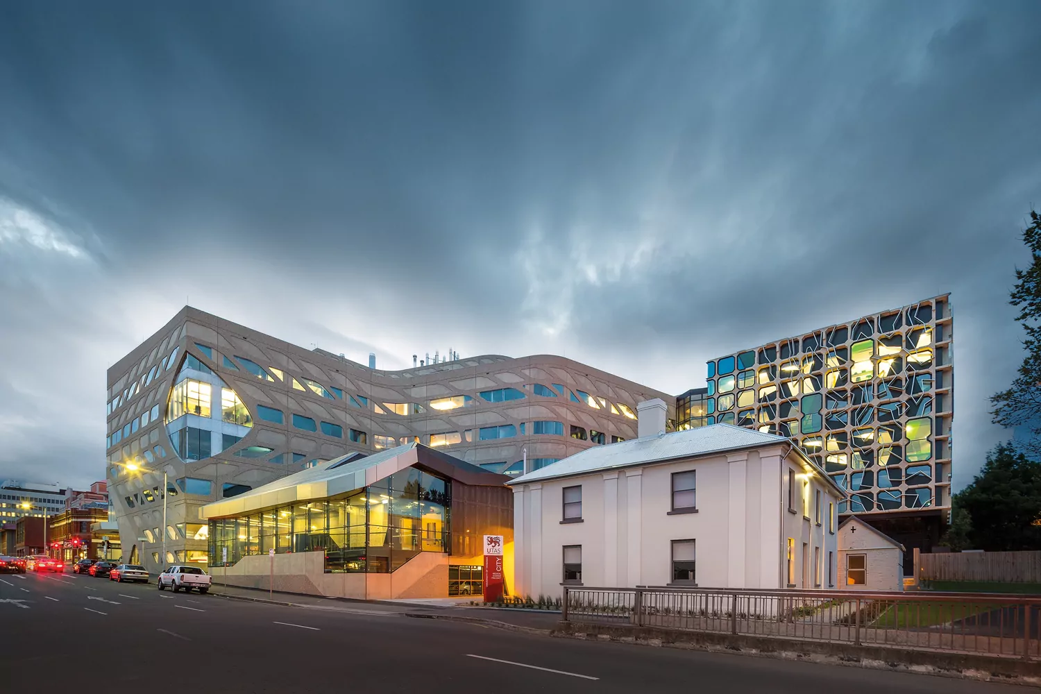 The iconic design of the University of Tasmania's Medical Sciences 2 building. The building is illuminated against an dark, overcast sky. The Medical Sciences 1 and Advocate House are visible in the foreground. The most eye-catching feature of the photo is the concrete lattice shading frame across the building, contour form and steel and coloured glazing of the building's façade.