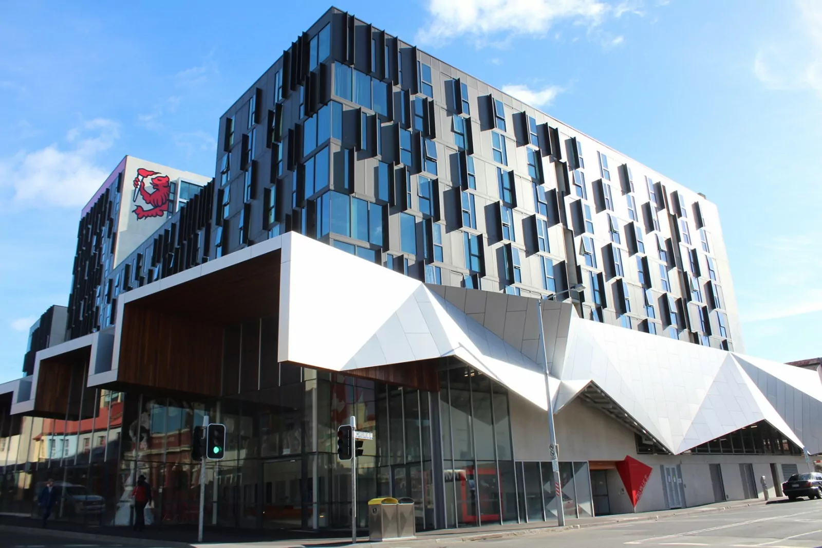A corner photo of the 442-unit, 4 towers of the National Rental Affordable Scheme accommodation at Melville Street. On one of the buildings, there is a prominent red lion logo of UTAS. The windows and external façade of fins and canopy reflect a bright sun.