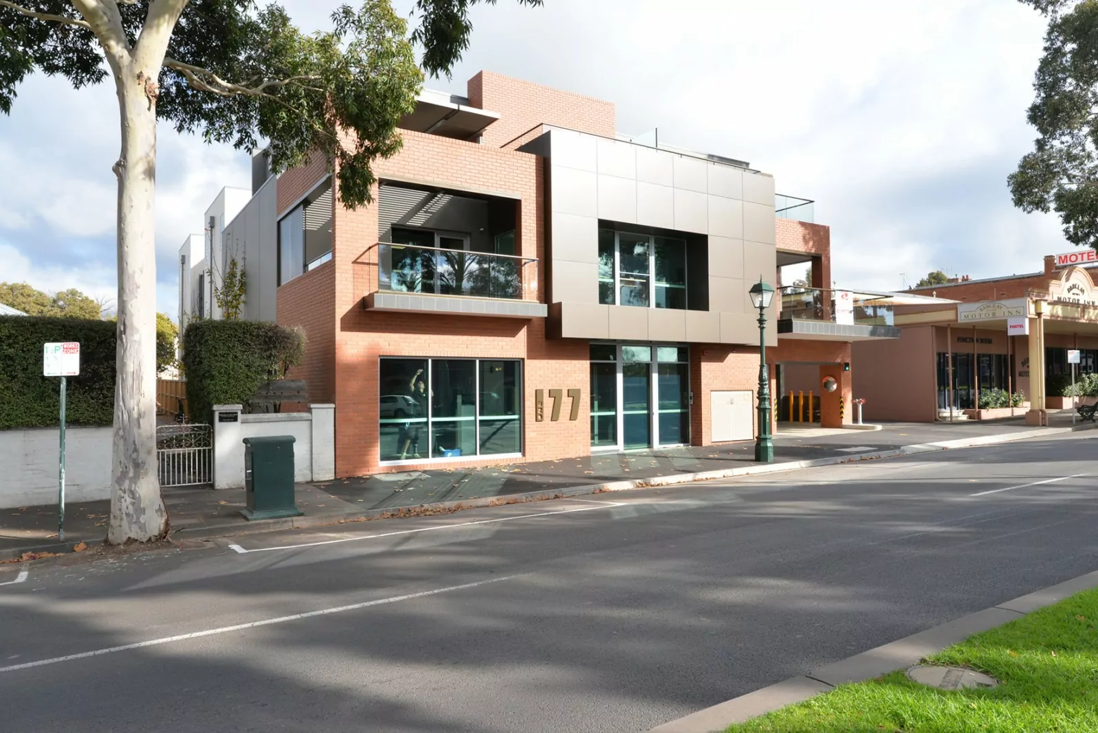 A modern three-story building with a red brick facade, grey cladding and stylised bronze metal One77 building logo. Inside the building someone is cleaning a window. Along the street, there are tall trees, a lamp post, neatly-cut hedges and street parking.