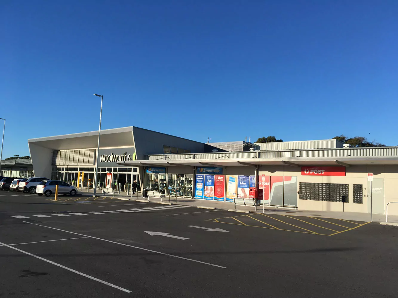 The retail development at Port Sorell. A wide road with marked yellow loading bays offers transit and parking for an Australia Post, Woolworths and other stores.