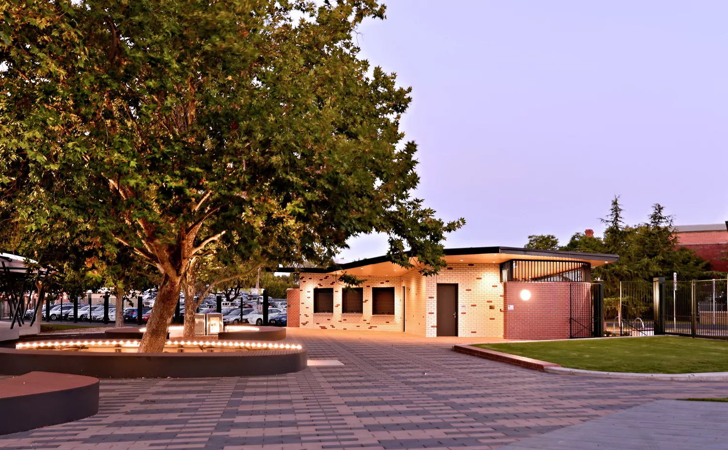 The Queen Elizabeth Oval kiosk and amenities building with lights turned on, lighting the building facade and outdoor tree timber lighting. The building is a mixture of red and glazed white bricks. Behind the building is a densely populated car parking.