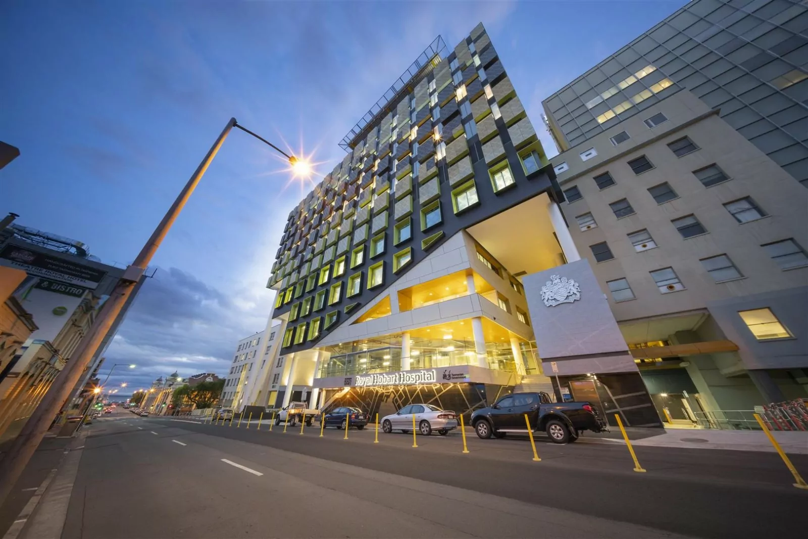 The redeveloped illuminated Royal Hobart Hospital, the largest Tasmanian capital infrastructure project to date. The facade features a crest, the bright logo of the hospital and a variety of greens, greys and yellows panelling around the windows.