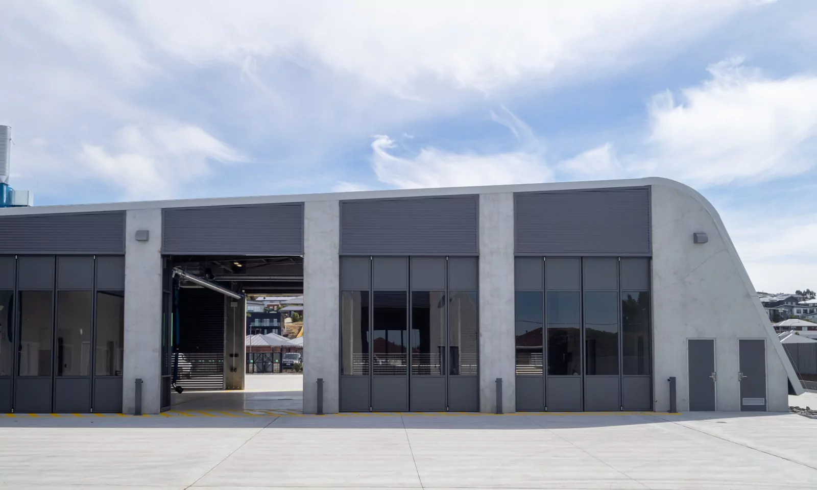A side-profile of the purpose-built Sorell Emergency Services Hub. The building has tall, specialised bi-folding doors to the fire engine bays leading outwards to an expansive concrete area. The surrounding area functions as a large parking, maneuvering space and washing bay area.