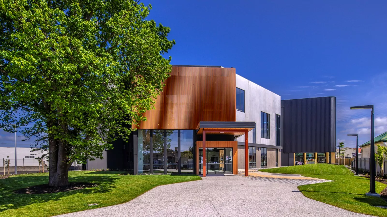 The front entrance of the Ulverstone Cultural Precinct ('Hive') building. A tall street stands alongside a building in the sunlight. Timber look aluminum battens provide sun shade. Grass and the pepper concrete footpath are reflected in the panels of glass windows.
