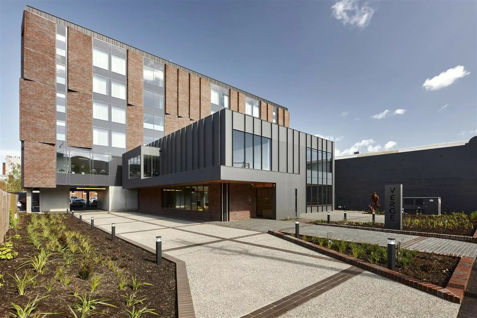 The external facade of the six-story boutique Hotel Verge in the Launceston CBD. A "Verge" logo is visible between plots of shrubs. The building itself is a pleasant mixture of brickwork and concrete.