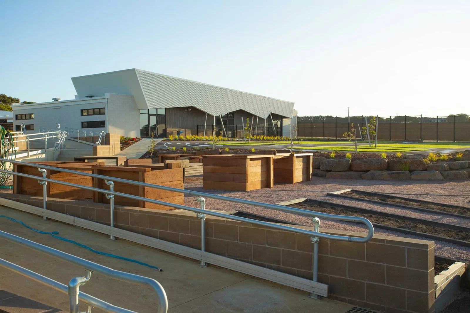 The Warrnambool Special Development School with its outdoor play areas. An accessible ramp leads a set of stairs. The surrounding area is landscaped and turfed with stone and timber feature.