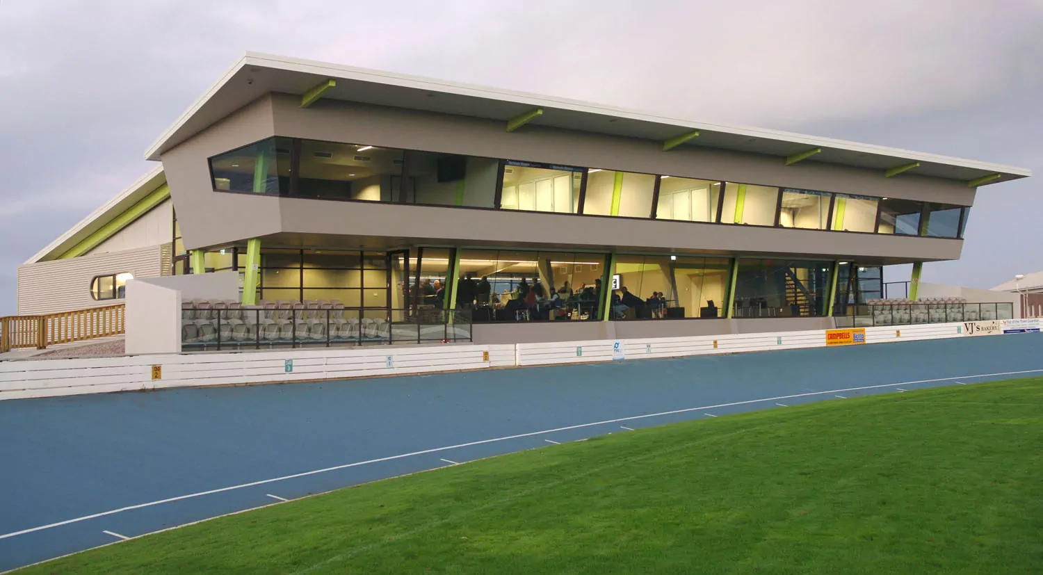 The modern and stylish West Park Sports Facility and Function Centre overlooks a sports oval and track. The silhouettes of a large group of people are visible through the windows.