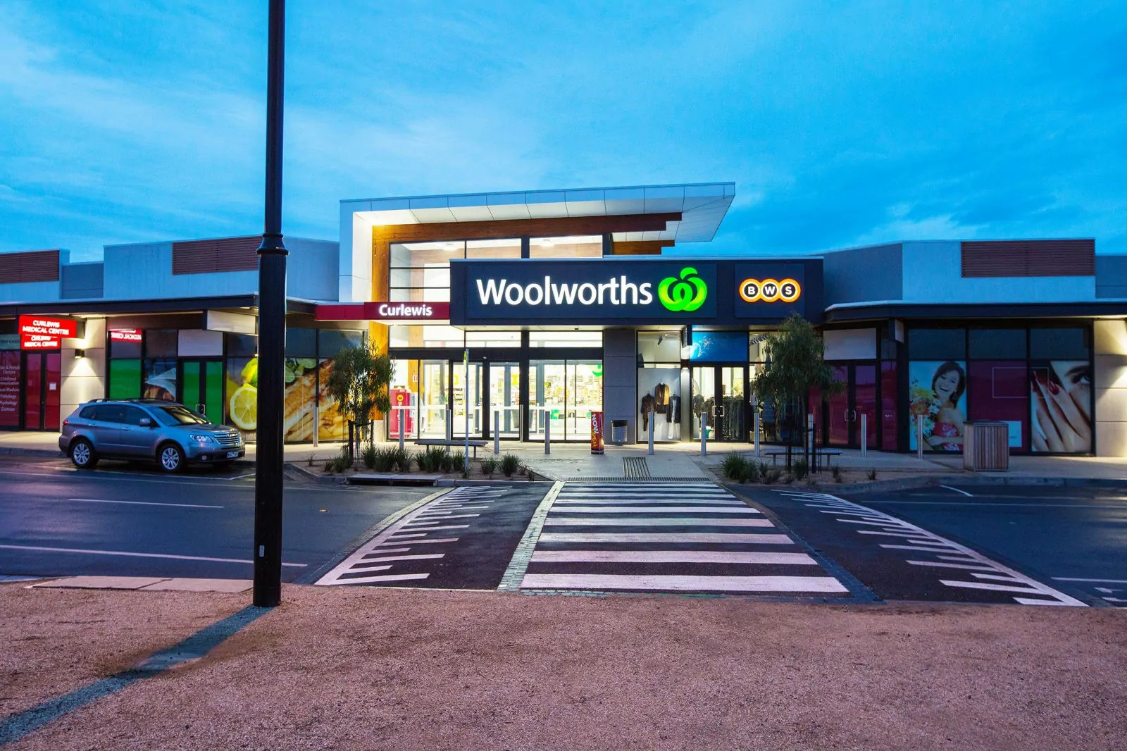 The iconic Woolworths logo is illuminated on the side of the Curlewis commercial centre. There is a parked car outside of the building.