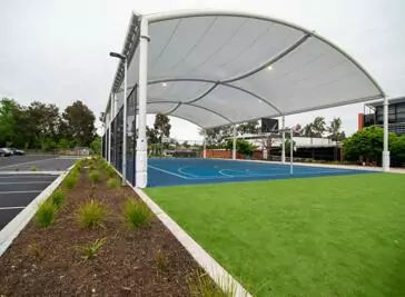 The sheltered outdoor sports court at Bendigo Senior Secondary College. The sides of the court are fenced off by a high metal fence, with car parking and surrounding strip of grass and native plants.