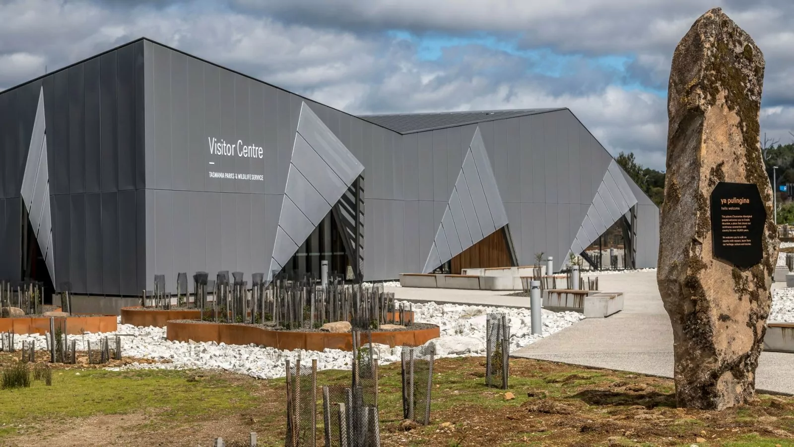 An external shot of the Cradle Mountain Gateway building with its concrete walkway. In the foreground is a plaque with information and traditional palawa kani greeting of "hello, welcome".