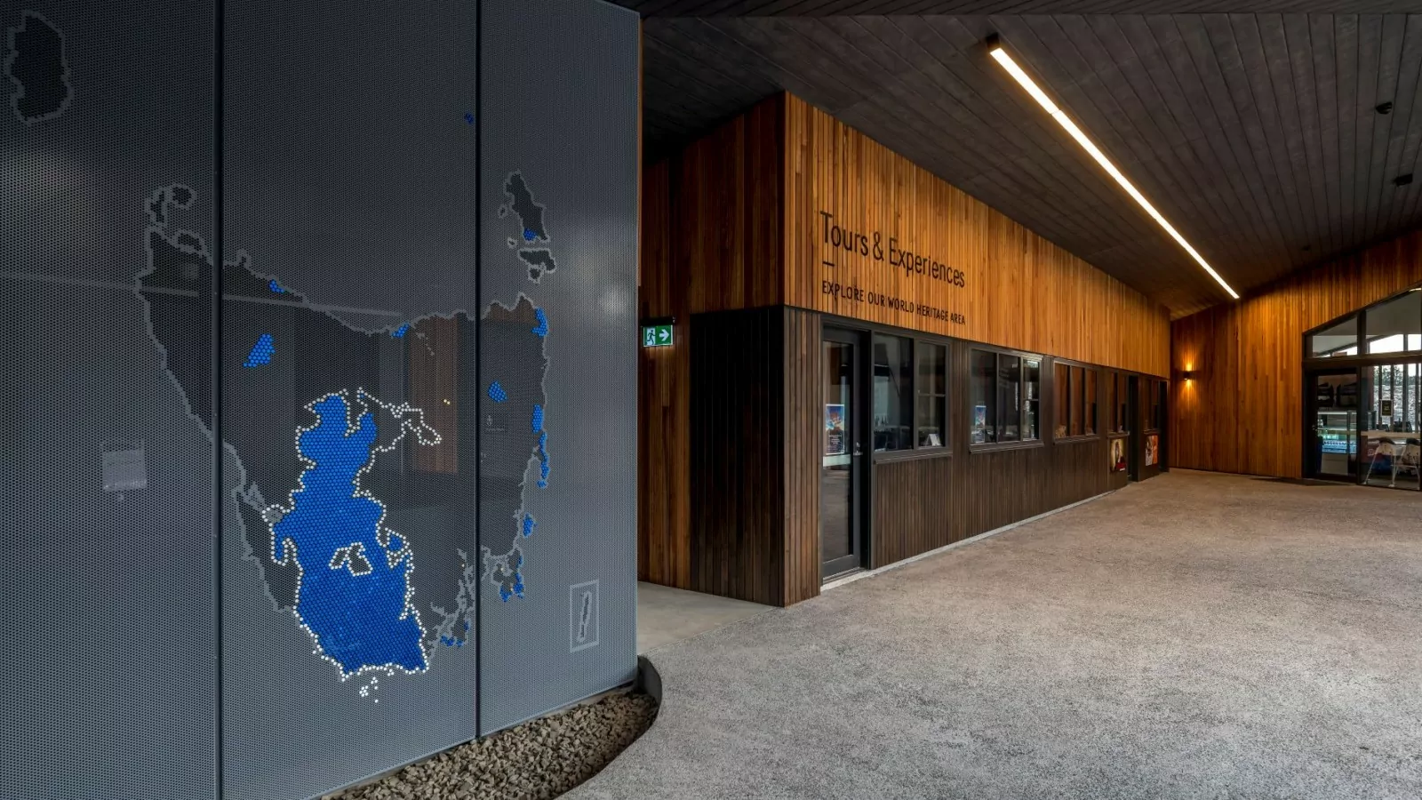 There is a map of Tasmania and its surrounding islands painted on perforated metal to left of a corridor and entrance to the "Tours and Experiences" section of the Cradle Mountain Gateway.