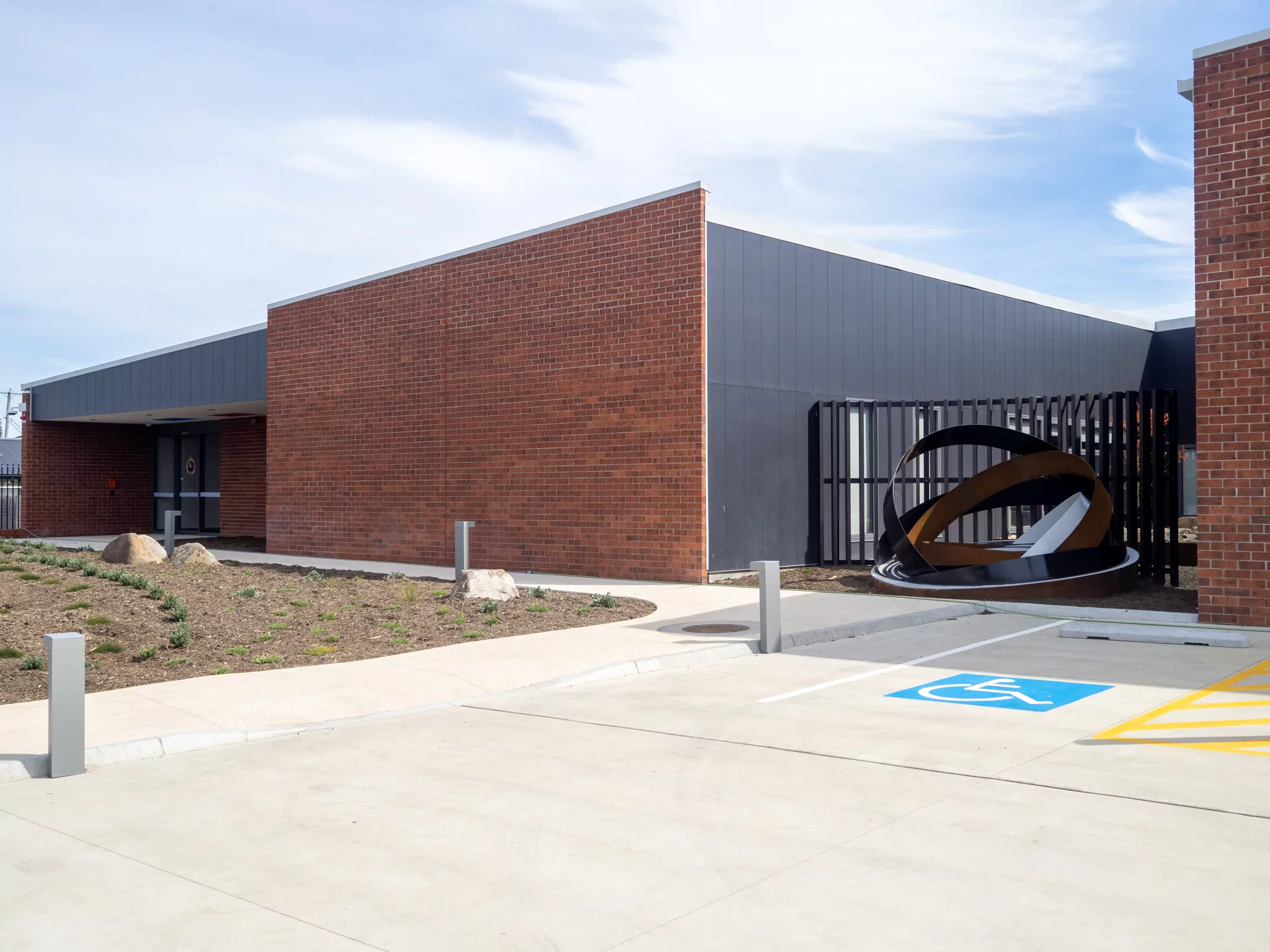 A concrete pedestrian walkway intersects with parking for people with disabilities, a semi-spherical statue and brickwall of the Sorell Emergency Services Hub.