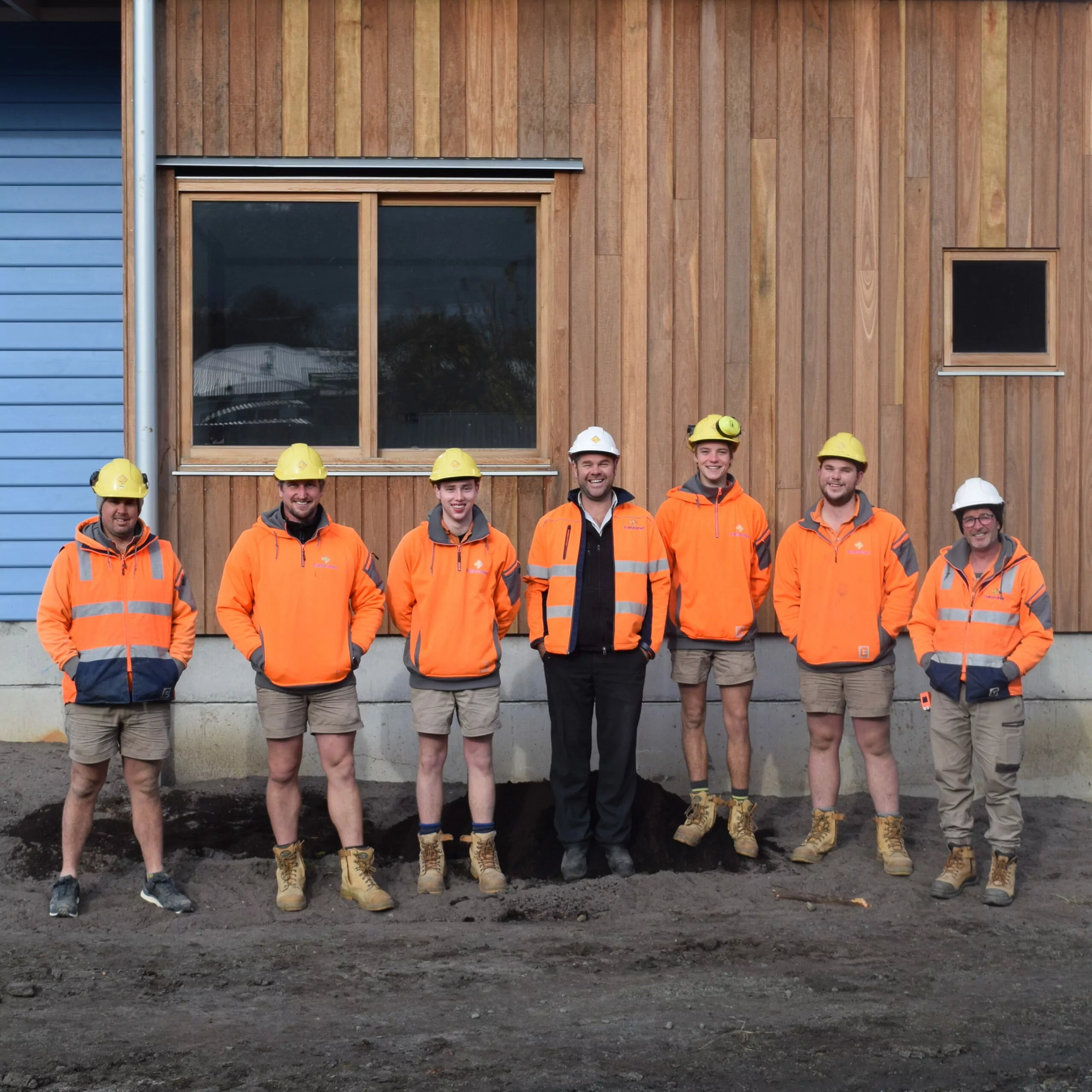 Seven construction workers in high vis clothing and hard hats standing on an active construction site. The wall they're standing in front of is of a building with a natural timber and blue weatherboard facade.