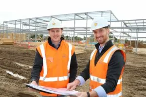 Two male staff members smile at the camera and gesture to held document. The pair are wearing hard hats and high vis jackets on a muddy worksite.