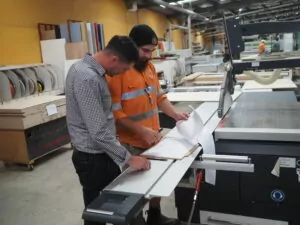 Two Fairbrother employees read through a clipboard of papers in the joinery workshop. One of the employees wears a grey and white patterned shirt and the other is in a Fairbrother branded high-vis shirt.