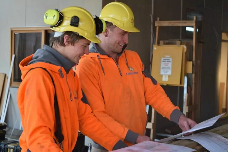 Two tradespeople smile and look at documents. They are wearing Fairbrother branded high vis hoodies and distinctive yellow hard hats.