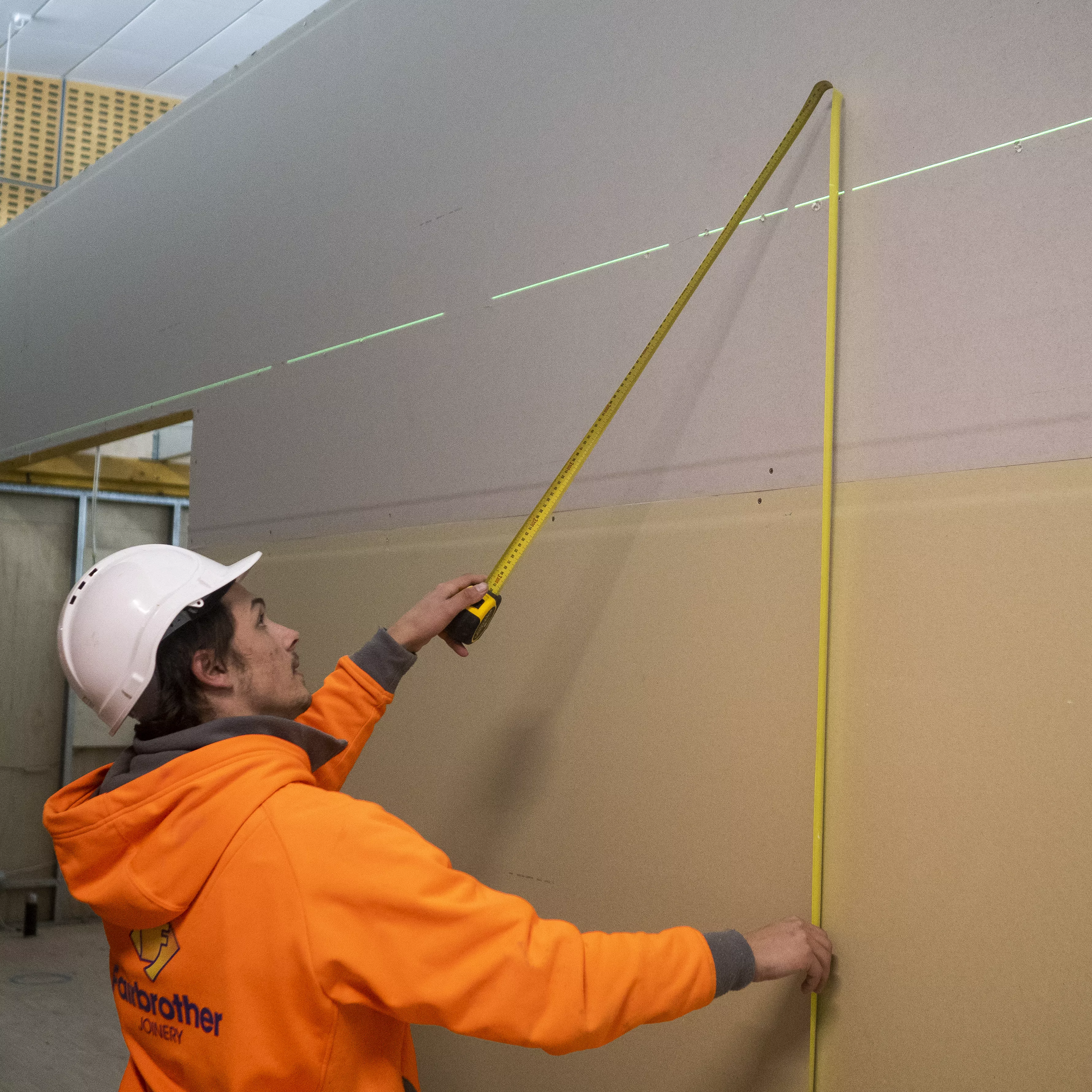 A fairbrother tradesperson with a high vis hoodie and white hard hat holds up a tape measure up against a concrete wall.