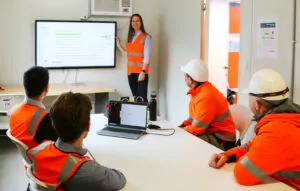 A female Fairbrother employee leads a presentation in an office-setting, in front of 4 other seated staff members. All of the people are wearing high vis clothing and other safety gear.